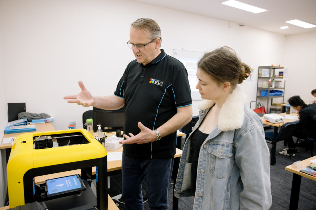 An Industry Skills Training instructor in a black and blue polo shirt is gesturing towards a bright yellow 3D printer, explaining its functions to a focused female student wearing a denim jacket with a cream collar. They are in a classroom setting with other students working in the background.