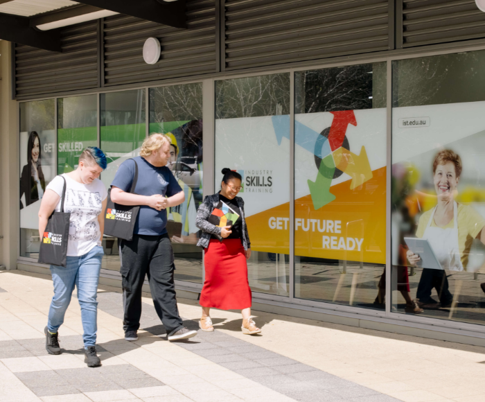 Three students are walking outside the Industry Skills Training Armadale campus, engrossed in conversation. They're passing by the campus's glass facade, which displays vibrant signage with phrases like 'GET SKILLED' and 'GET FUTURE READY'. One student is holding a notebook and another is carrying a tote bag, indicating active participation in campus life.