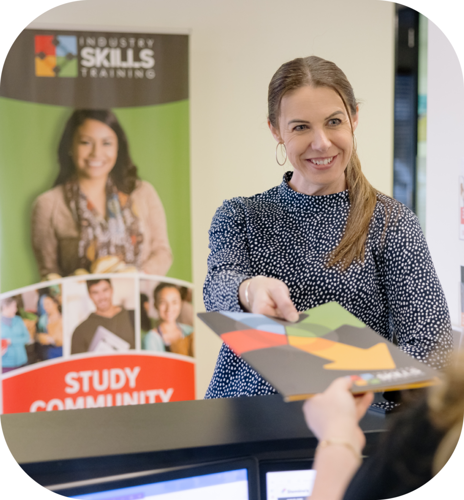 An engaging staff member at Industry Skills Training, who appears friendly and professional, is in the process of handing over documents at the reception area. The background is adorned with vibrant educational banners, reflecting the dynamic learning community at the institution.