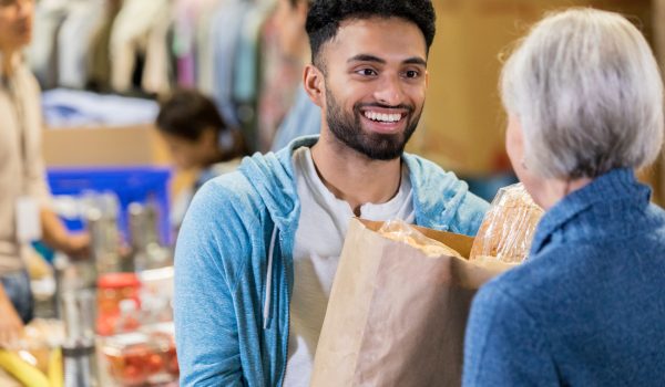 Smiling male food bank volunteer gives a senior woman a paper bag filled with groceries.