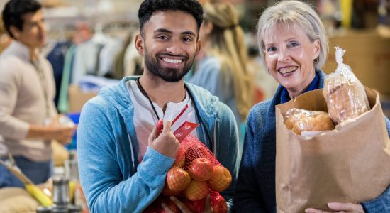 A young male food bank volunteer stands next to a senior female customer.  They hold groceries as they smile for the camera.