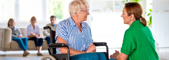 Friendly home nurse talking with senior woman in retirement house. Elderly lady sitting in wheelchair.