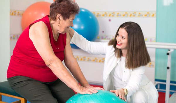 Old woman exercising with fitness ball on the physical therapy with help of the physical therapist in the hospital.